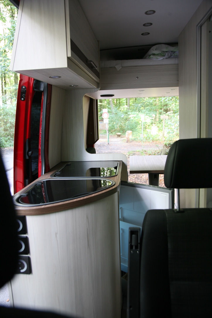 Innenraum des Sprinter-Campers mit Küchenzeile, Spülbecken, Kochfeld, geöffnetem Kühlschrank und Hubbett im oberen Bereich